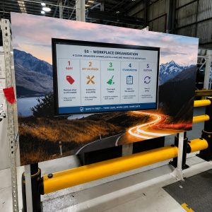 Large indoor banner showing a 5S workplace organisation poster (Sort, Set in Order, Shine, Standardise, Sustain) with icons, mounted in an industrial setting near yellow safety rails.
