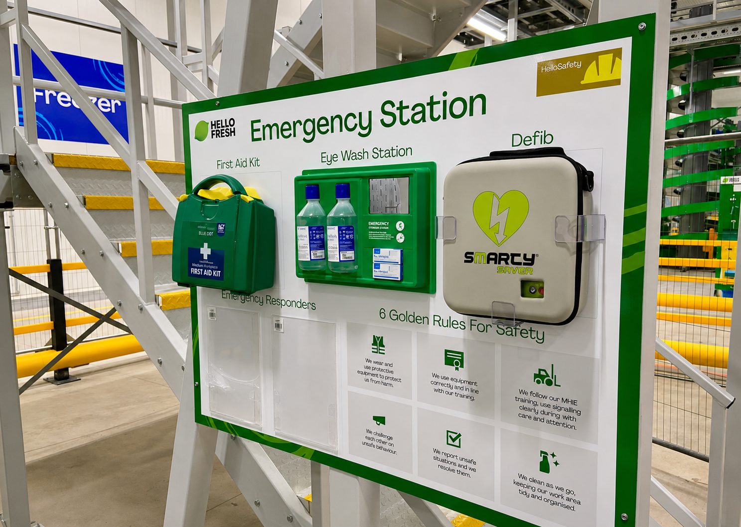 Industrial Emergency Station board featuring a First Aid Kit, Eye Wash Station, and Defibrillator case mounted on a metal framework in a workspace.