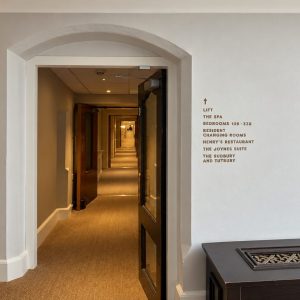 Hotel hallway seen through an arched doorway, with a potted plant on the left, wall light, and framed floral art on the right.
