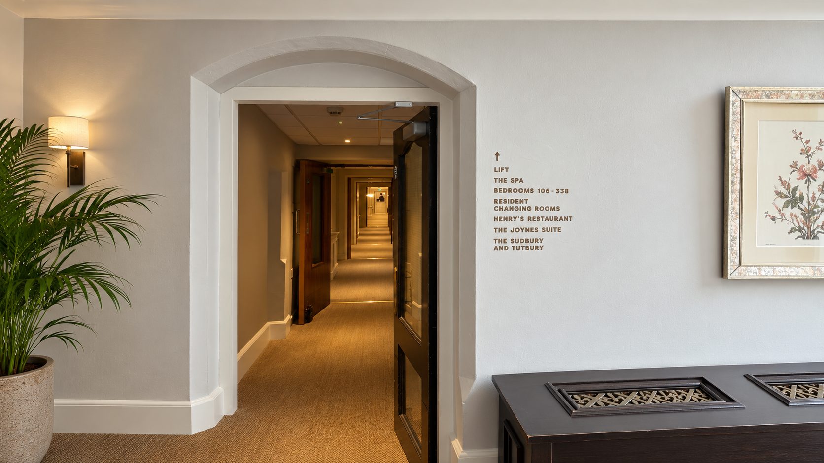 Hotel hallway seen through an arched doorway, with a potted plant on the left, wall light, and framed floral art on the right.