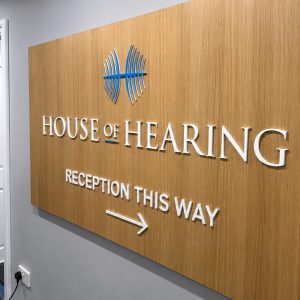 Wood-paneled sign reading 'HOUSE OF HEARING' with a blue logo and 'RECEPTION THIS WAY' arrow.