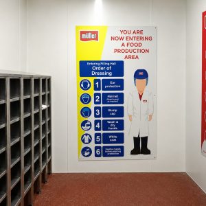 Signage in a food production area displaying dressing order: ear protection, hairnet, bump cap, wash hands, white coat beside a cartoon worker; yellow and blue panels with steps.