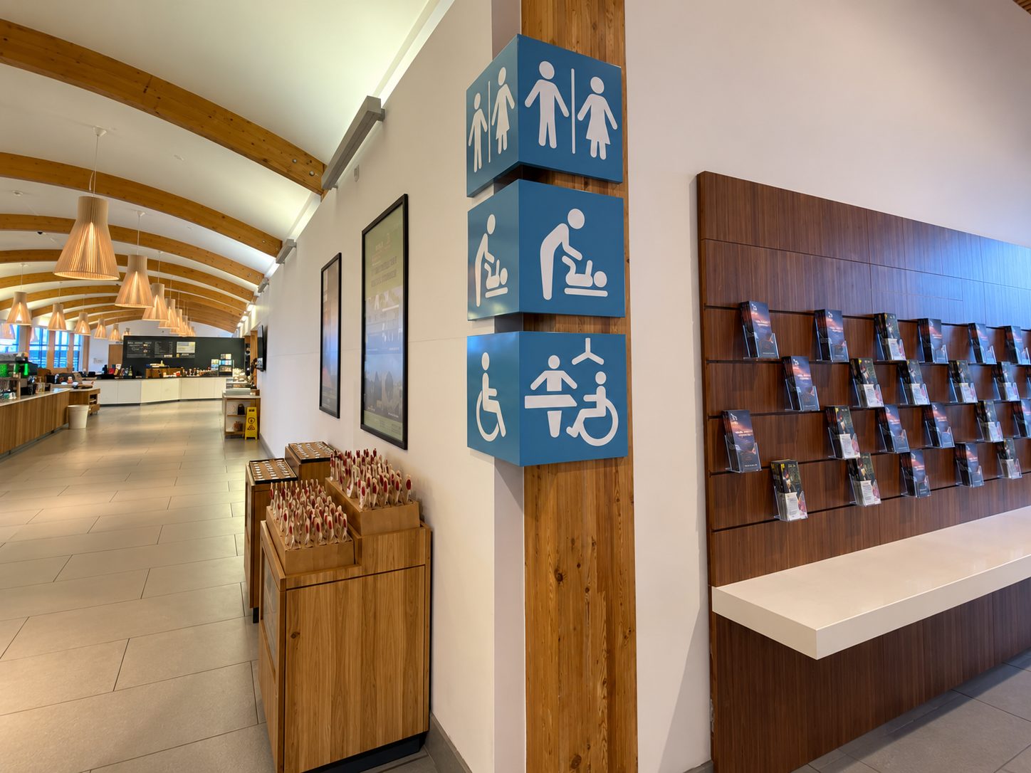Interior cafe or lounge with wooden arched ceiling, warm pendant lights, and a magazine rack along a wood-paneled wall near blue accessibility sign cubes on a pillar