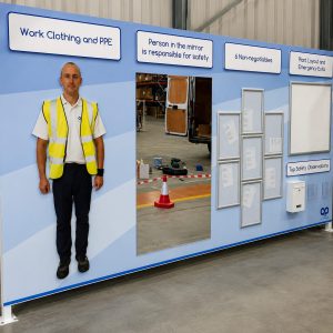Worker in a high-visibility vest stands beside a safety display wall in a warehouse.
