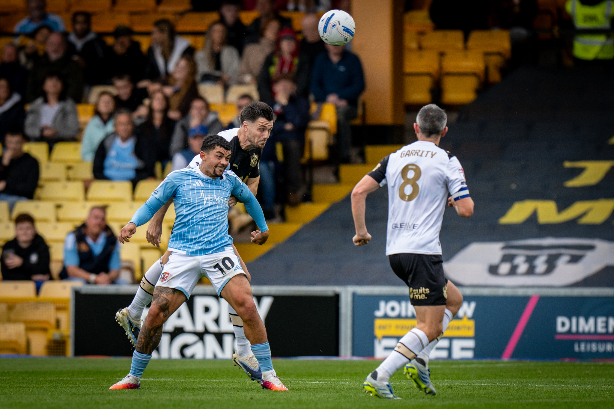 Two soccer players leap to head the ball during a match, one in light blue kit and white shorts, the other in a dark kit, with yellow stands behind them.