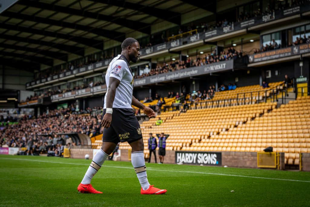 Soccer player in white jersey walks along a stadium pitch with green grass and yellow seats in the background.