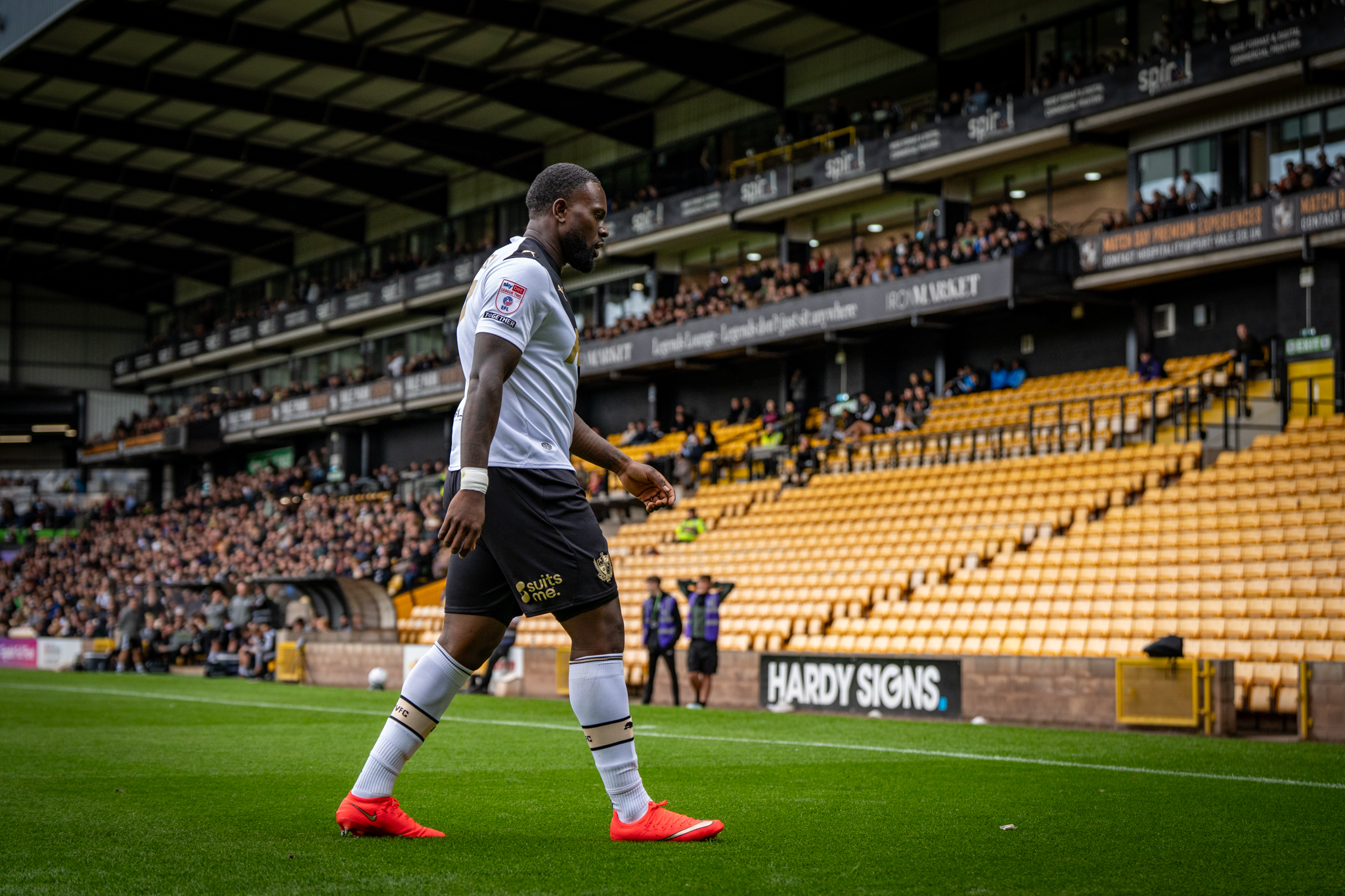 Soccer player in white jersey walks along a stadium pitch with green grass and yellow seats in the background.
