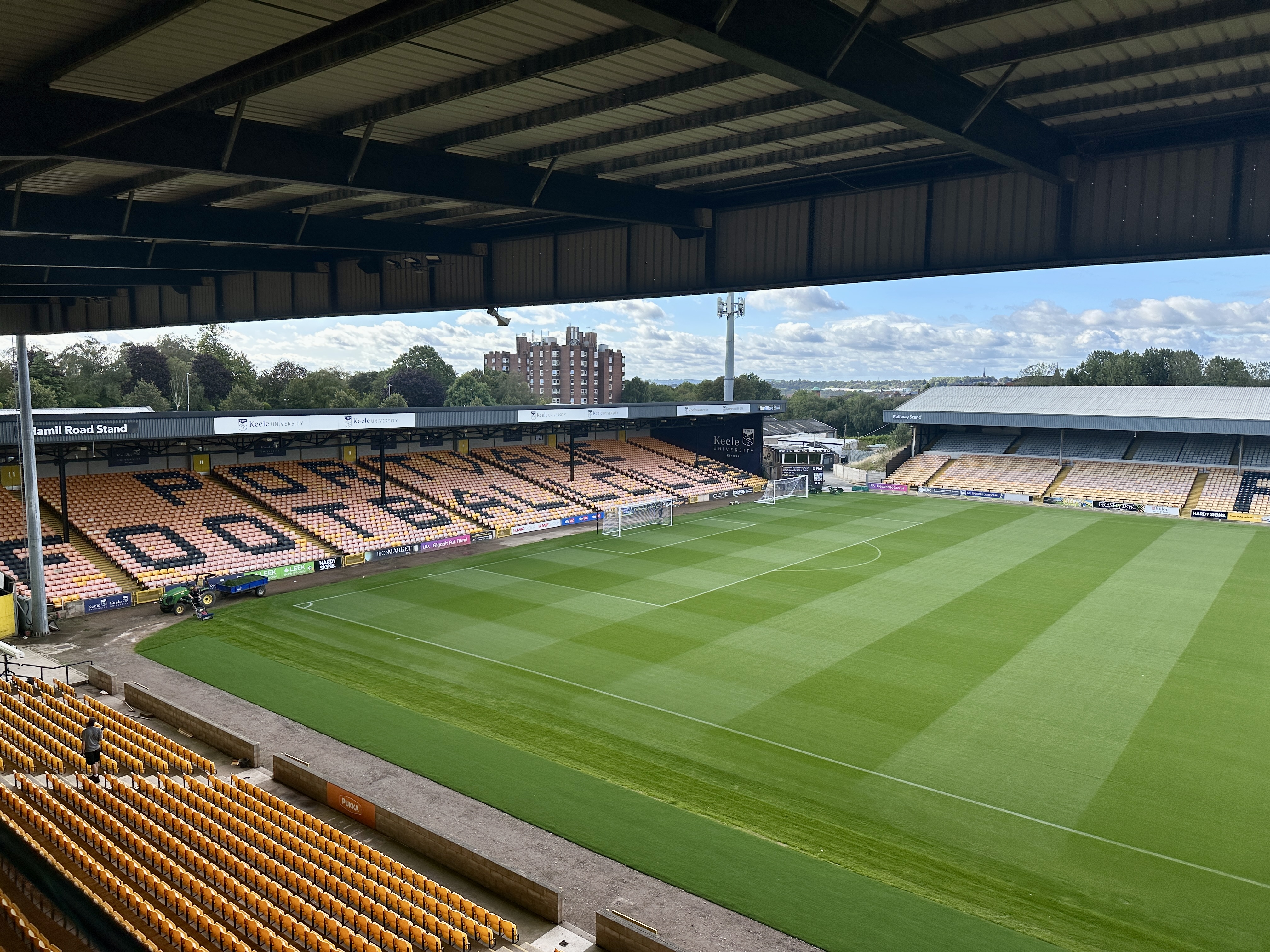 Empty orange-seated football stadium with a green pitch, viewed from the upper grandstand under a dark metal roof on a sunny day.