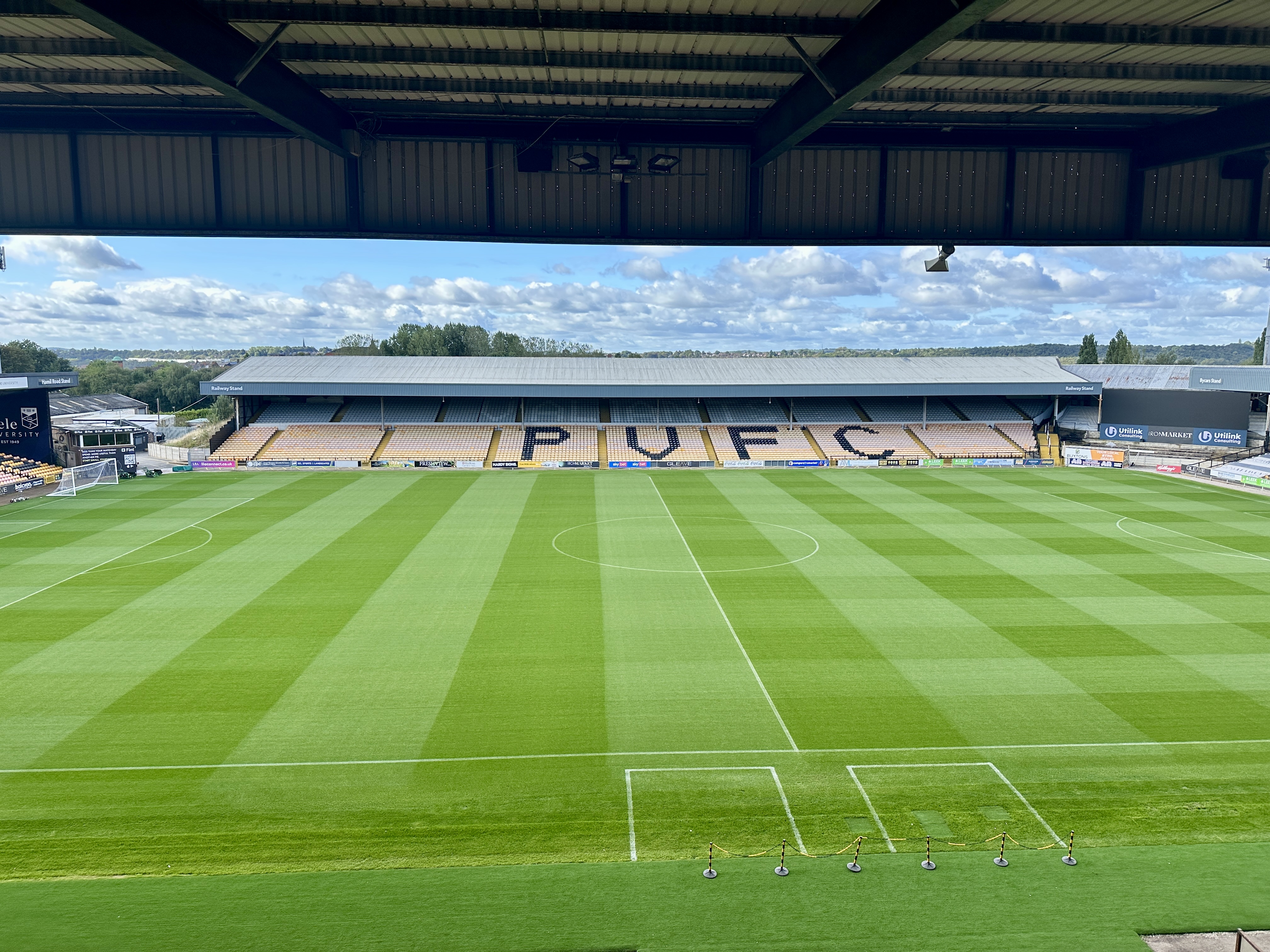 Wide view of a soccer stadium: manicured green pitch with striped patterns, empty yellow seating behind a long stands roof.