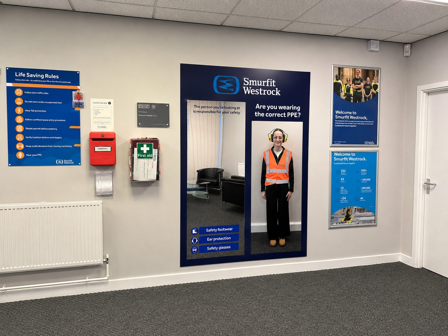 Person in a bright orange safety vest stands in a doorway cutout labeled Smurfit Westrock, with PPE posters nearby.