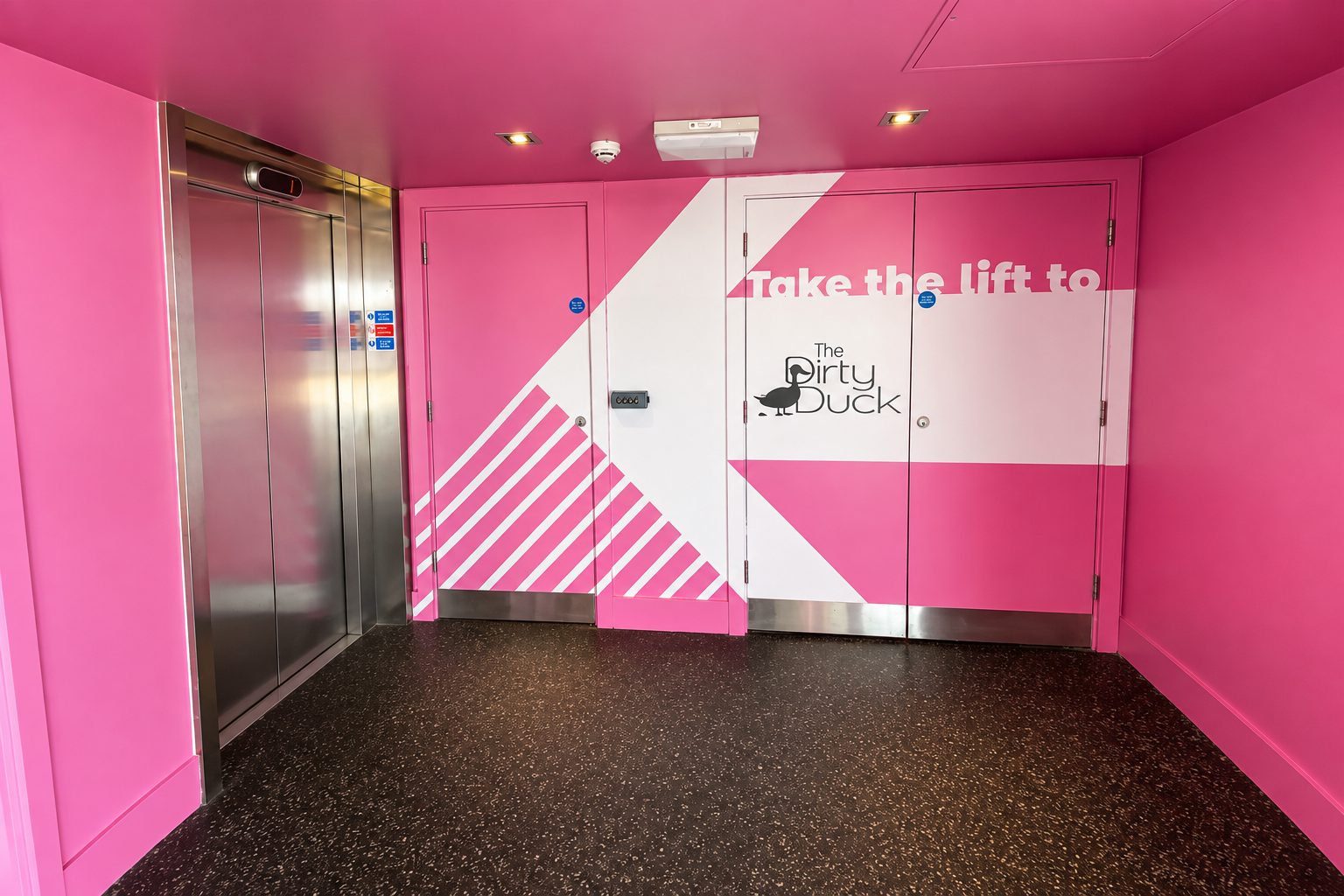 Pink-walled hallway with an elevator on the left and a door labeled The Dirty Duck; text on wall reads 'Take the lift to' and doors feature white and pink geometric design.