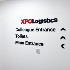 Wall sign for XPO Logistics directing to Colleague Entrance, Toilets, and Main Entrance with three black arrows (up, up, left) on the right side of the text.