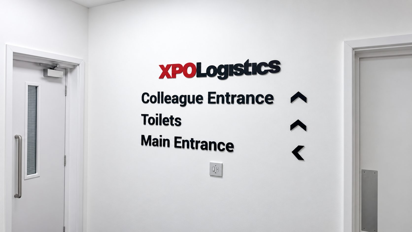 Wall sign for XPO Logistics directing to Colleague Entrance, Toilets, and Main Entrance with three black arrows (up, up, left) on the right side of the text.